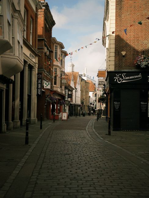 A narrow, cobbled pedestrian street in a town centre with buildings on both sides featuring retail shops and cafes. The buildings have a mix of brick and plaster facades, with some displaying hanging flower baskets and signage. Overhead, a string of small triangular flags is suspended across the street, fluttering gently in the breeze. The street curves slightly into the distance, leading towards a white domed structure visible in the background. The lighting suggests a clear day with partial sunlight casting soft shadows on the pavement. On the right side, there is a closed black shop with a rolled-down shutter, while other shops along the street display open entrances and window displays. Modern black bollards are positioned along the edges of the pavement, providing separation from the roadway, which is free of traffic. This scene exemplifies usual urban shopping district environments, potentially prepared for local crowd support or public events, often subject to occasional cleanup or rubbish collection by local services such as Rubbish Clearance Uxbridge to maintain cleanliness in shared commercial areas.