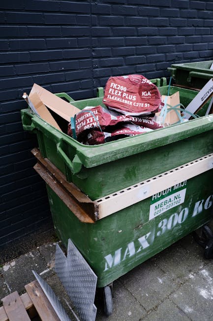 A green wheeled rubbish skip filled with black bags of construction or garden waste, placed against a dark brick wall outdoors on a paved surface. The bags feature red and white markings and text, indicating they contain flexible groundwork materials. The skip has a white branding strip on its side, with some minor debris visible on the ground nearby. The scene appears to be in an outdoor commercial or residential area where private waste collection services, such as Rubbish Clearance Uxbridge, might be employed for waste removal or site clearance. The skip is partially overflowing, with bags stacked on top, and the environment is lit by natural daylight, emphasizing the textures of the waste bags and the sturdy green plastic of the skip, supported by a metal frame and wheels for mobility.