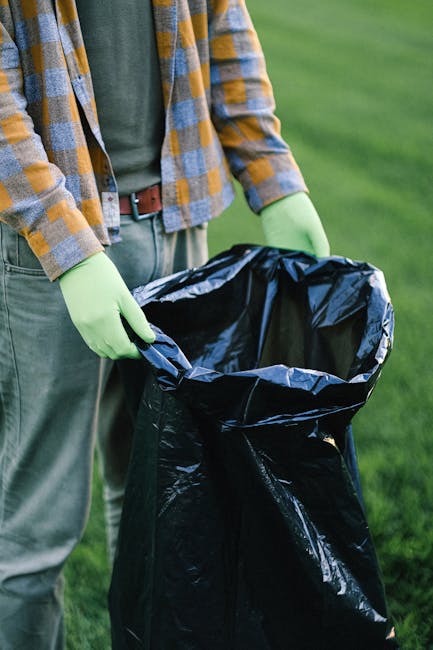 A person wearing a grey and yellow checkered shirt, with a green long-sleeved top underneath, is holding open the top of a large black rubbish bag lined with a shiny black plastic liner. The individual is wearing light green gloves for handling waste. The scene is set outdoors on a green grassy area, with the person's lower torso and arms visible, but their face is not shown. In the background, blurred green foliage and grass suggest a park or garden setting. The image emphasizes the process of waste collection or rubbish disposal, which relates to private waste handling services provided by Rubbish Clearance Uxbridge, showcasing the preparation for rubbish removal. The overall lighting appears natural, highlighting the textures of the bag, gloves, and clothing, consistent with an outdoor environment suitable for rubbish clearance activities.