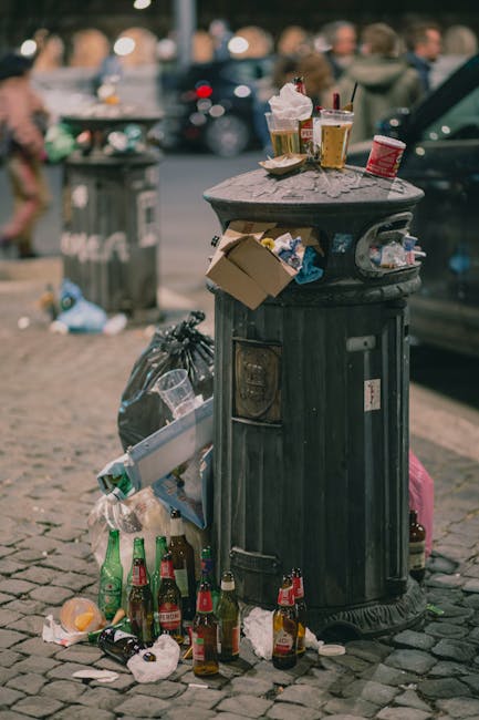 A collection of black rubbish bags stacked against the exterior wall of a building on a small concrete pavement in an urban setting. The building has a textured green wooden façade with peeling paint and red trim around the windows and door. A set of wooden stairs with a dark finish leads up to a white door at the top, which has a small overhang with red and white striped trim. To the left, there is a door with graffiti and a small metal electrical box attached to the wall, with a power cable hanging below it. In the background, a street with parked cars and a multi-storey brick building is visible under overcast skies, indicating a typical city environment. The scene suggests a private rubbish collection or on-site clearance, operated by Rubbish Clearance Uxbridge, situated within a residential or commercial area that requires waste removal services. The overall image emphasizes the practical aspects of rubbish disposal outside a property, with the bags and urban context highlighting the need for professional waste management solutions.