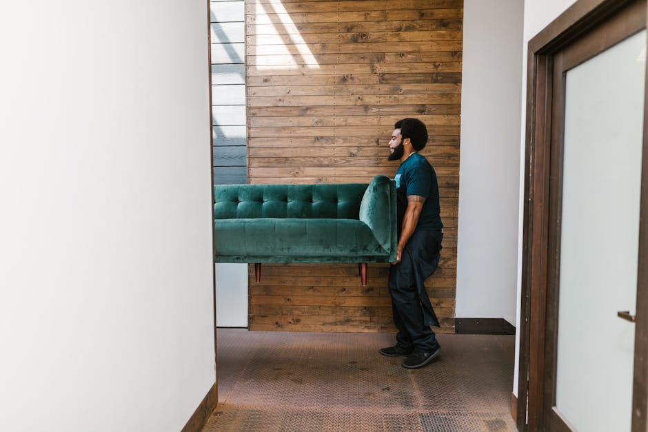 A man with a dark beard, wearing a teal uniform top and black pants, is shown lifting a green upholstered sofa with a button-tufted design and wooden legs. The sofa appears to have a velvet-like texture and is positioned vertically in a narrow indoor space, with the backrest facing a wooden wall made of horizontally fitted planks in warm brown tones. The flooring is covered with a dark brown textured carpet. To the right, there is a door with a dark wooden frame and frosted glass panel. The setting suggests an interior room or hallway, possibly during a rubbish removal or furniture clearance service, with natural light illuminating the scene. The overall environment appears clean and organized, consistent with professional waste management or furniture collection activities by Rubbish Clearance Uxbridge.