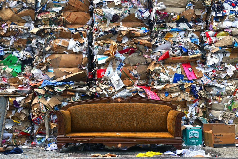 A vintage brown upholstered sofa with ornate wooden framing sits on a pavement in front of a large, overflowing pile of mixed waste and shredded paper, including brown cardboard boxes, crumpled paper pieces, and various packaging materials. The waste extends vertically, creating a dense, chaotic backdrop that appears to be plastic and paper waste, some of which shows printed and colored surfaces. The scene is outdoors, with natural ambient lighting emphasizing the texture and disorder of the waste while the sofa remains relatively intact amidst the clutter. To the right, there are some cardboard boxes labeled with brands, and placing the scene in context of rubbish removal services, it suggests a comparison with alternative waste disposal methods such as private rubbish clearance by Rubbish Clearance Uxbridge, highlighting the challenge of managing bulky waste accumulation in urban settings.
